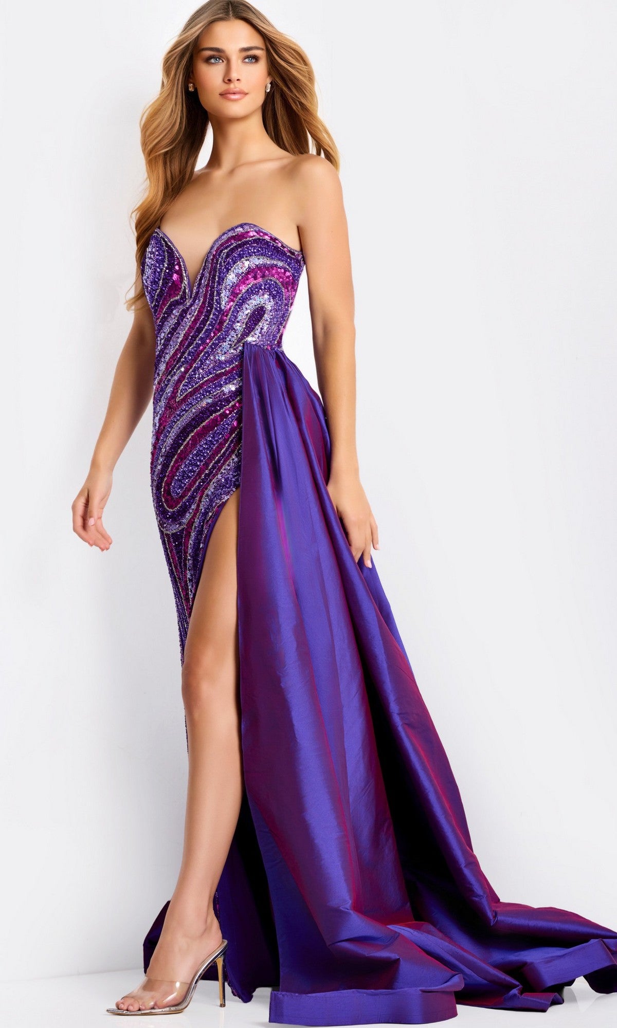A woman confidently wears the Jovani 49226, a strapless purple long prom dress with multicolored sequins, thigh-high slit, detachable dramatic overskirt, and clear high-heeled sandals.